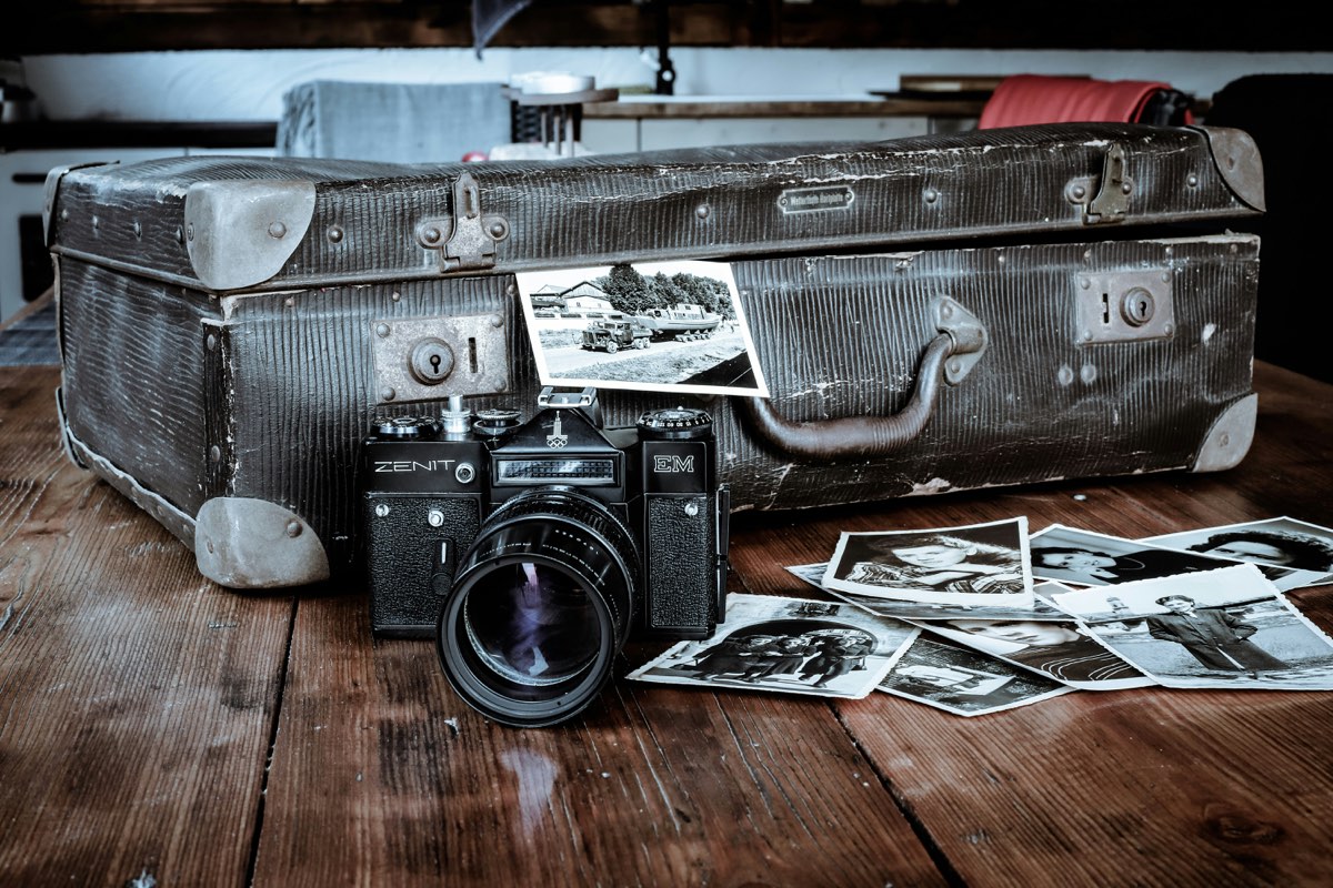 An old camera and a battered suitcase on a wooden table, with a scattered heap of black and white photographs beside them